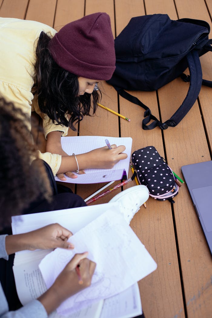 High angle of crop anonymous diverse schoolchildren writing in workbook while doing homework on wooden platform on street